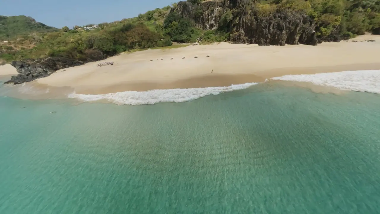 Aerial shot skimming turquoise water toward an empty beach below green cliffs.
