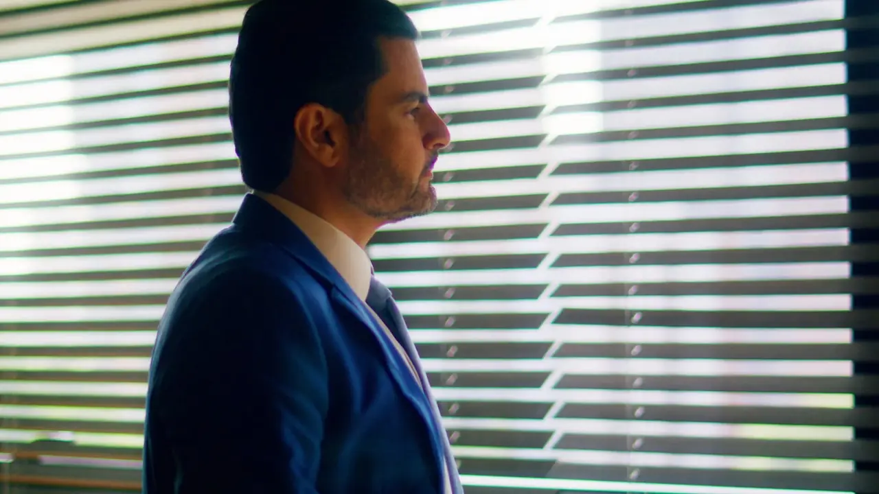 A lawyer in a blue suit stands in profile against sunlit venetian blinds.